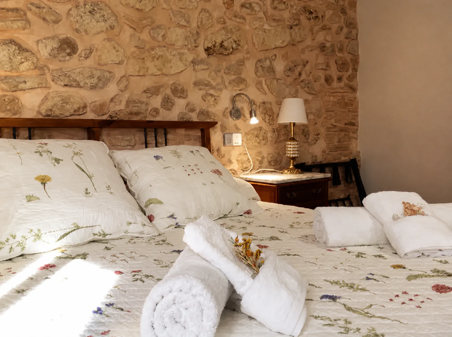 Cama doble de la habitación L'Alzina con colcha de flores, toallas y pared de piedra.